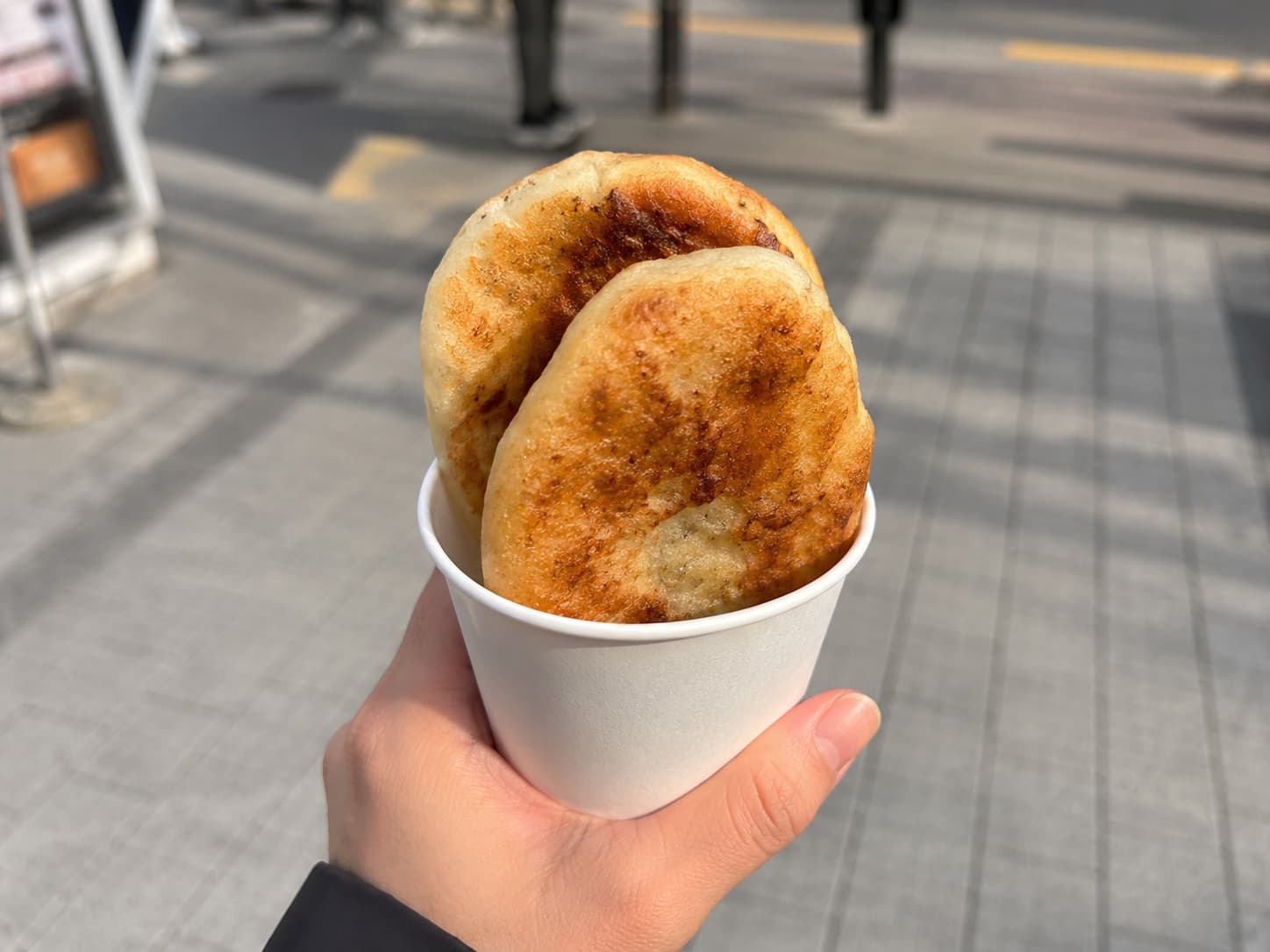 A hand holding a white paper cup with two whole, golden-brown hotteok on a Korean street.